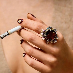 Könnte beinhalten: Eine Hand mit dunklem Nagellack und weißen Blumenakzenten hält eine brennende Zigarette. Ein großer, verzierter Ring mit einem dunkelroten Edelstein und floralen Details ziert den Ringfinger. Der Ring hat goldene und blaue Akzente.