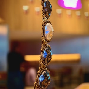 May include: A gold-toned bracelet with oval-shaped, iridescent stones. The bracelet features ornate, filigree-style metalwork around each stone. The stones alternate between a pearly white and a dark, reflective color, creating a striking contrast.