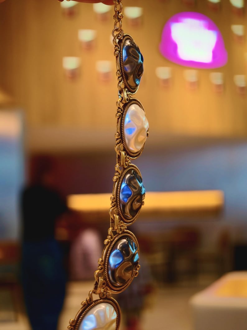 May include: A gold-toned bracelet with oval-shaped, iridescent stones. The bracelet features ornate, filigree-style metalwork around each stone. The stones alternate between a pearly white and a dark, reflective color, creating a striking contrast.
