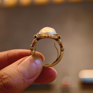 May include: A gold ring featuring a large, luminous pearl. The ring has an ornate, vintage design with intricate detailing around the pearl setting and band. The ring is held up, showcasing its elegant craftsmanship.