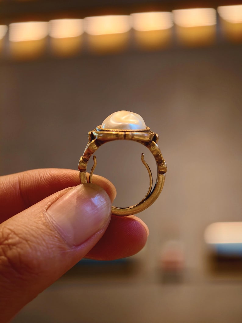 May include: A gold ring featuring a large, luminous pearl. The ring has an ornate, vintage design with intricate detailing around the pearl setting and band. The ring is held up, showcasing its elegant craftsmanship.