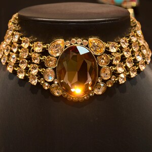 Pu&ograve; includere: Una collana dorata con una grande gemma ovale color ambra al centro. La collana &egrave; adornata da numerosi cristalli pi&ugrave; piccoli, rotondi e a goccia, color ambra, che creano un design scintillante e lussuoso. La collana &egrave; esposta su un manichino nero.