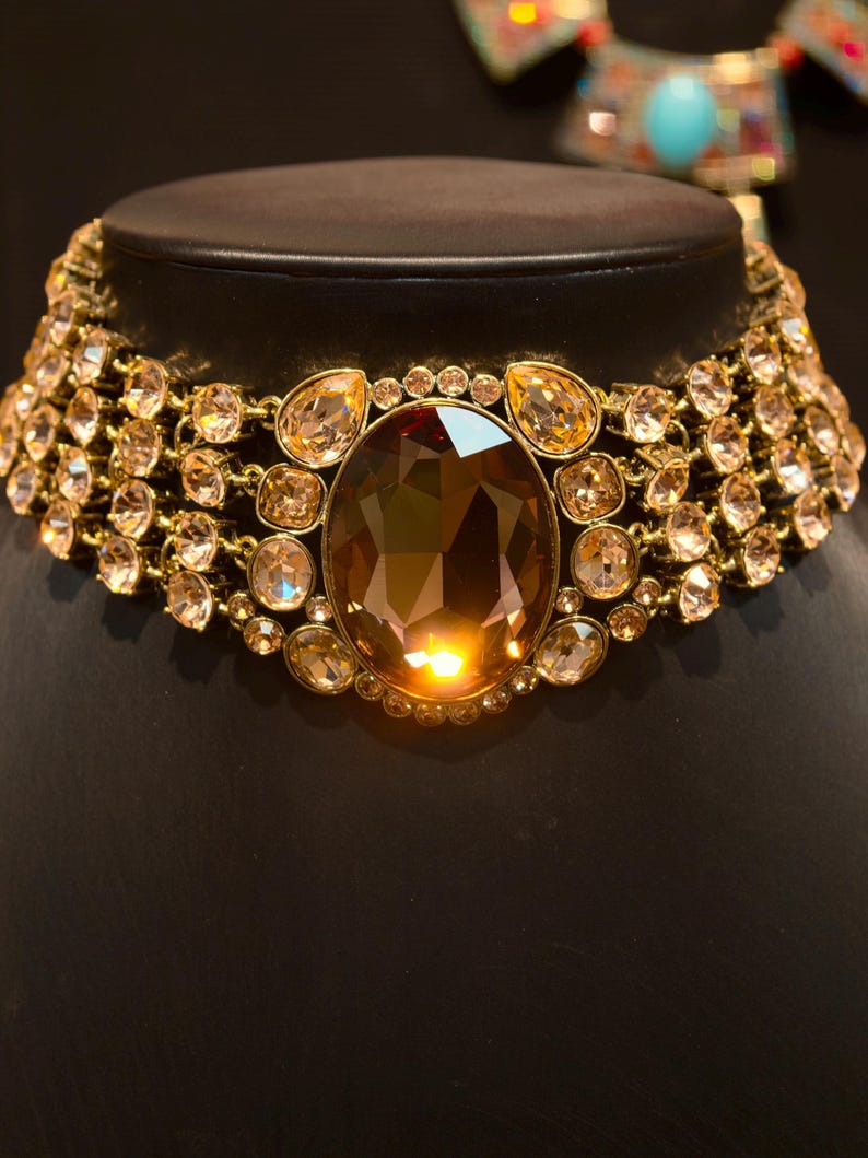Pu&ograve; includere: Una collana dorata con una grande gemma ovale color ambra al centro. La collana &egrave; adornata da numerosi cristalli pi&ugrave; piccoli, rotondi e a goccia, color ambra, che creano un design scintillante e lussuoso. La collana &egrave; esposta su un manichino nero.