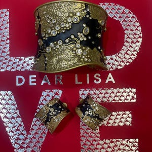 May include: A gold-toned cuff bracelet and matching earrings with black enamel and clear crystal accents. The jewelry is set against a red background with the words "DEAR LISA LOVE" in silver lettering.