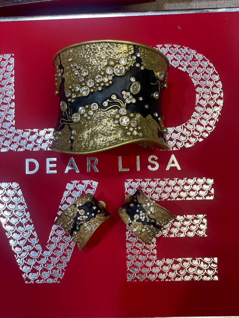 May include: A gold-toned cuff bracelet and matching earrings with black enamel and clear crystal accents. The jewelry is set against a red background with the words "DEAR LISA LOVE" in silver lettering.