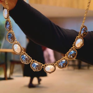 May include: A gold-toned necklace featuring oval-shaped pendants with alternating white and blue-grey centers. The necklace has an ornate, vintage-inspired design, with a delicate chain and decorative frames around each pendant.