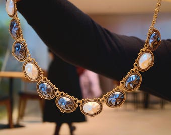Op de afbeelding: Een goudkleurige ketting met ovale hangers met afwisselend witte en blauwgrijze centra. De ketting heeft een sierlijk, vintage-geïnspireerd ontwerp, met een delicate ketting en decoratieve frames rond elke hanger.