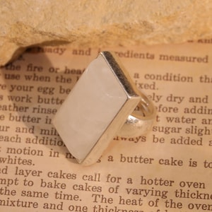 May include: A silver ring with a large, rectangular, pale-colored stone. The stone is set in a silver band. The ring is displayed on a vintage book page with text. The ring's design is simple and elegant.