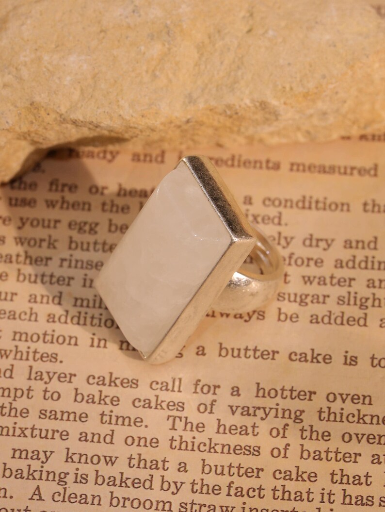 May include: A silver ring with a large, rectangular, pale-colored stone. The stone is set in a silver band. The ring is displayed on a vintage book page with text. The ring's design is simple and elegant.