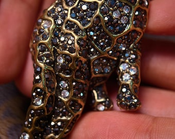 May include: A detailed, jeweled leopard figurine. The sculpture features a gold-toned metal frame filled with dark and clear crystals. The leopard's eyes are a bright green. The figurine is held in a hand.