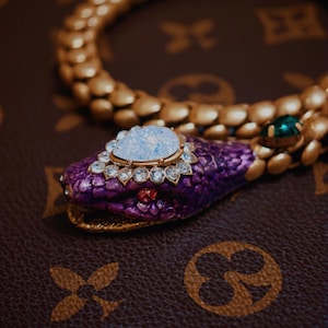 Puede incluir: Un collar de serpiente en tono dorado con un cuerpo texturizado similar a escamas. La cabeza de la serpiente tiene un acabado en esmalte morado intenso con una gran piedra iridiscente en forma de pera, rodeada de cristales claros y rojos.