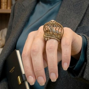 May include: A gold-toned crown ring with intricate detailing and small gemstones. The ring is worn on a finger, showcasing its ornate design. The ring's crown shape is the focal point, suggesting a regal or vintage aesthetic.