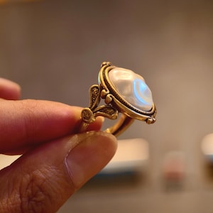 May include: A gold-toned ring featuring a large, pearly white, oval-shaped stone. The ring has an ornate, vintage-inspired design with delicate filigree details and a decorative setting. The ring is held between fingers, showcasing its elegant craftsmanship.