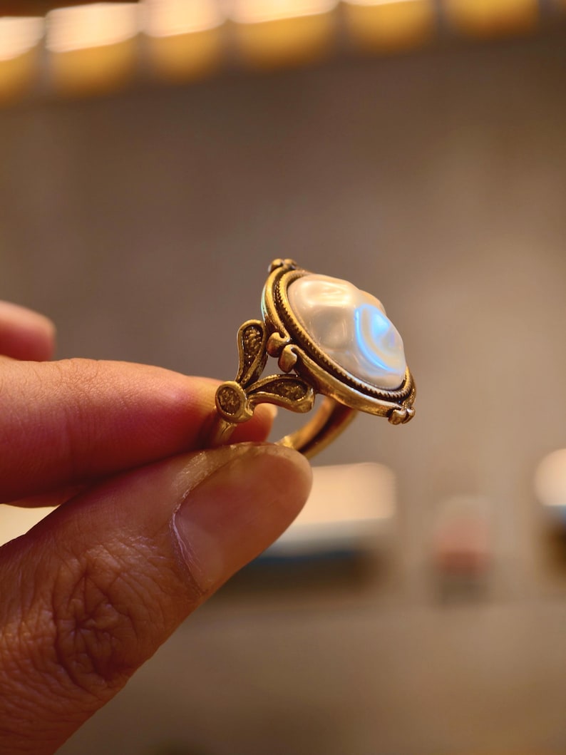 May include: A gold-toned ring featuring a large, pearly white, oval-shaped stone. The ring has an ornate, vintage-inspired design with delicate filigree details and a decorative setting. The ring is held between fingers, showcasing its elegant craftsmanship.