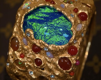 Op de afbeelding: Een goudkleurige manchetarmband met een getextureerd oppervlak. De armband heeft een grote, centrale blauwe en groene steen, omringd door kleinere rode, bruine en iriserende stenen. Kleine heldere stenen zijn ook in het goud ingebed.
