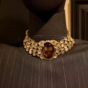 Pu&ograve; includere: Una collana dorata con una grande gemma ovale marrone al centro. La collana &egrave; adornata con cristalli pi&ugrave; piccoli, trasparenti e color ambra. La collana &egrave; esposta su un manichino nero.