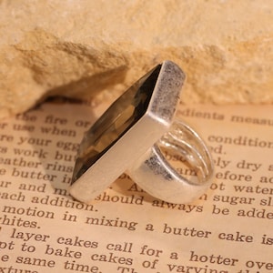 May include: A silver ring with a large, rectangular, smoky quartz gemstone. The ring has a wide band and a modern, geometric design. The gemstone is a translucent brown color. The ring is set against a vintage paper background.