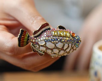 May include: A decorative fish-shaped trinket box, adorned with a multitude of colourful rhinestones and gems. The fish's body is primarily white with a scale-like texture, while the fins and tail display intricate details. The gems are in shades of pink, yellow, blue, and orange.