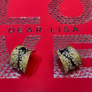 May include: Two gold-toned cuff bracelets with black enamel accents and clear crystal embellishments. The bracelets are displayed on a red surface with the words "DEAR LISA" in silver lettering.