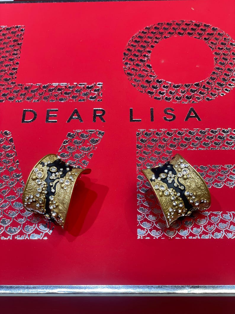 May include: Two gold-toned cuff bracelets with black enamel accents and clear crystal embellishments. The bracelets are displayed on a red surface with the words "DEAR LISA" in silver lettering.