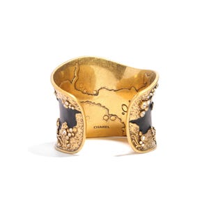 May include: Gold-toned cuff bracelet with black enamel accents and sparkling crystal embellishments. The interior features a map design and the name "CHAREL". A statement piece of jewelry.