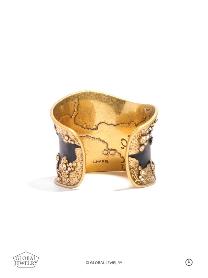 May include: Gold-toned cuff bracelet with black enamel accents and sparkling crystal embellishments. The interior features a map design and the name "CHAREL". A statement piece of jewelry.