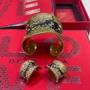 May include: A gold-toned cuff bracelet and matching earrings set with black enamel accents and sparkling clear crystals. The jewelry is displayed on a red surface with the text "DEAR LISA".
