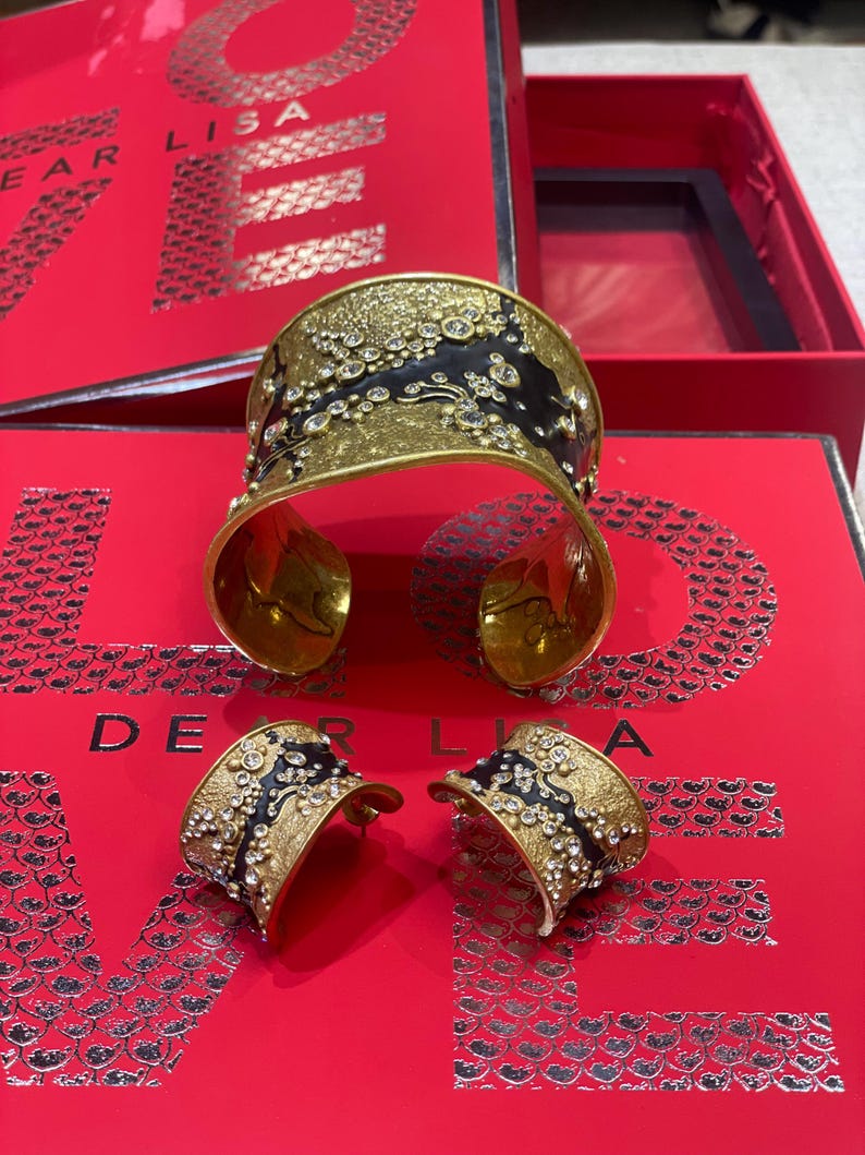 May include: A gold-toned cuff bracelet and matching earrings set with black enamel accents and sparkling clear crystals. The jewelry is displayed on a red surface with the text "DEAR LISA".
