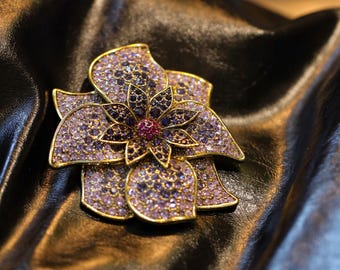 May include: A gold flower brooch, adorned with purple and dark purple gemstones. The brooch features a central red gemstone. It is set against a dark, textured backdrop.