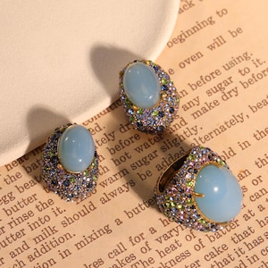 May include: Three pieces of jewelry: two earrings and a ring. Each piece features a large, oval, light blue stone surrounded by smaller, colorful gemstones. The ring has a gold-colored band. The jewelry is set on a vintage paper background.