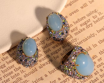 May include: Three pieces of jewelry: two earrings and a ring. Each piece features a large, oval, light blue stone surrounded by smaller, colorful gemstones. The ring has a gold-colored band. The jewelry is set on a vintage paper background.