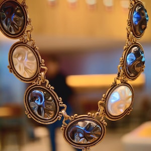 May include: A gold-toned necklace featuring oval-shaped pendants with iridescent and white accents. The pendants are linked together with ornate connectors, creating a statement piece. The necklace is displayed against a blurred background.