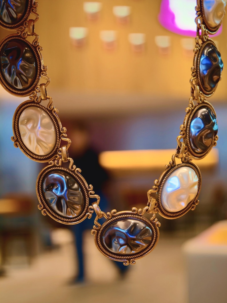 May include: A gold-toned necklace featuring oval-shaped pendants with iridescent and white accents. The pendants are linked together with ornate connectors, creating a statement piece. The necklace is displayed against a blurred background.