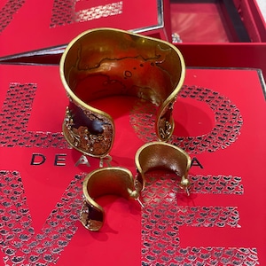 May include: A gold-toned cuff bracelet and matching hoop earrings with a dark burgundy enamel and crystal accents. The jewelry sits on a red surface with the words "DEAR LISA" printed in silver.