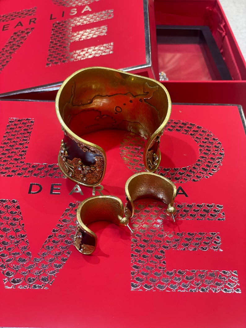 May include: A gold-toned cuff bracelet and matching hoop earrings with a dark burgundy enamel and crystal accents. The jewelry sits on a red surface with the words "DEAR LISA" printed in silver.