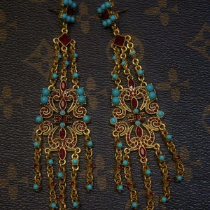 May include: A pair of ornate gold-toned earrings with red and turquoise accents. The earrings feature intricate filigree designs, dangling chains, and small turquoise beads. The earrings are set against a dark brown background.