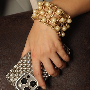 May include: A gold-toned bracelet with faux pearls and a silver-toned phone case with a grid pattern of small, round metal studs. The bracelet is worn on a wrist, and the phone is held in the hand.