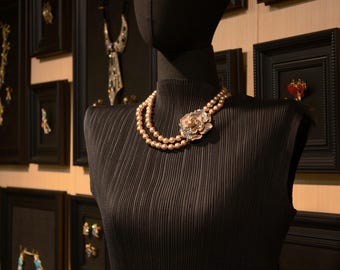 以下が含まれることがあります： 黒いマネキンが、花のブローチが付いた二連のパールネックレスを展示しています。 マネキンは、額入りのジュエリーが飾られた壁を背景にしており、アクセサリーのコレクションを紹介しています。