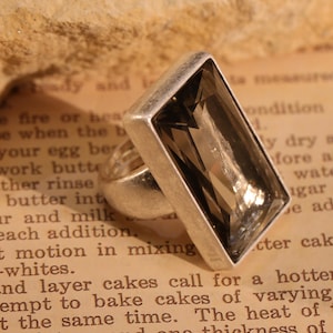 May include: A silver ring with a large, rectangular, smoky quartz gemstone. The ring is set against a vintage book page background, showcasing the ring's design and color.