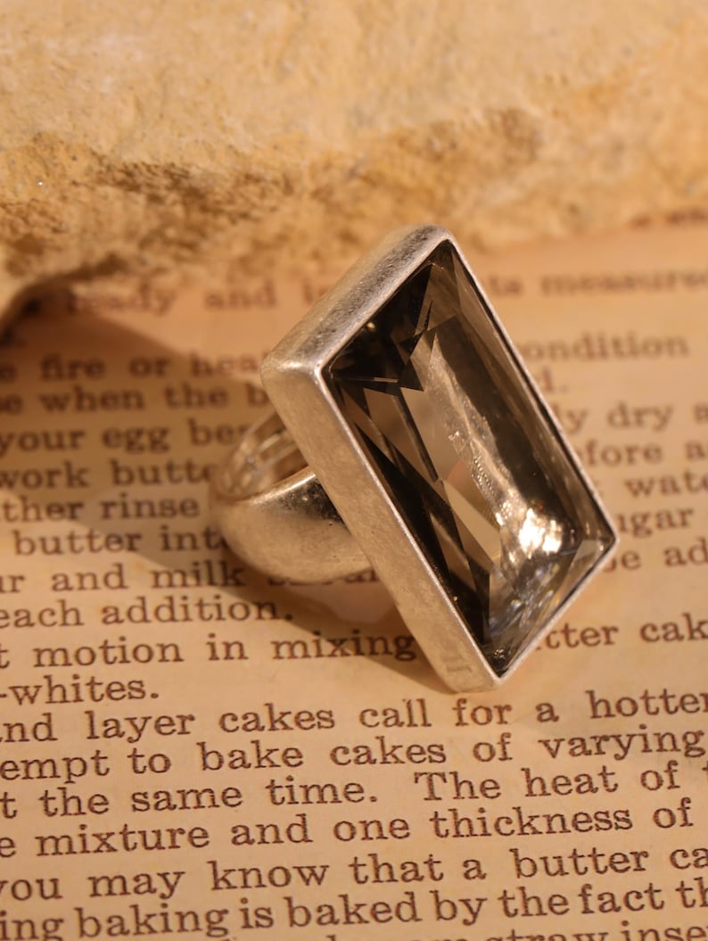 May include: A silver ring with a large, rectangular, smoky quartz gemstone. The ring is set against a vintage book page background, showcasing the ring's design and color.