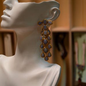 Op de afbeelding: Een paar goudkleurige oorhangers met lichtblauwe ovale en druppelvormige stenen. De oorbellen worden getoond op een witte mannequinhoofd, waarbij hun trapsgewijze ontwerp en elegante stijl worden benadrukt.