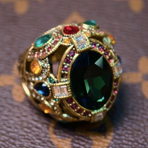 Könnte beinhalten: Ein goldfarbener Ring mit einem ovalen, dunkelgrünen Mittelstein, umgeben von kleineren, farbigen Edelsteinen. Der Ring weist aufwendige Details auf und ist vor einem braunen Hintergrund platziert.