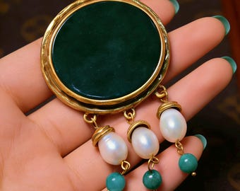 May include: A gold brooch with a large, round, dark green stone centre. Three dangling elements feature white pearls and teal beads. The brooch has a classic, elegant design.