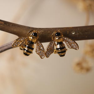May include: A pair of gold-coloured bee-shaped earrings. Each earring has a black and gold striped body, clear crystal-like wings, and a small black head. The earrings are displayed on a brown branch.