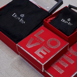 May include: Red gift boxes with silver trim and the word "LOVE" spelled out in silver glitter. The boxes contain black fabric pouches with the "Dear Lisa" logo. One box has a clear lid.