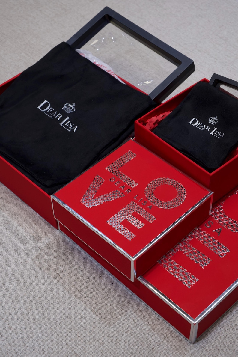 May include: Red gift boxes with silver trim and the word "LOVE" spelled out in silver glitter. The boxes contain black fabric pouches with the "Dear Lisa" logo. One box has a clear lid.