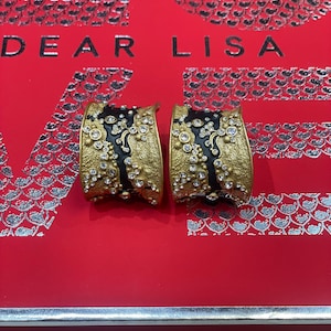 May include: A pair of gold-toned earrings with a black enamel center, adorned with clear crystals. The earrings are set against a red background with the text "DEAR LISA". The earrings have a textured finish and a curved design.