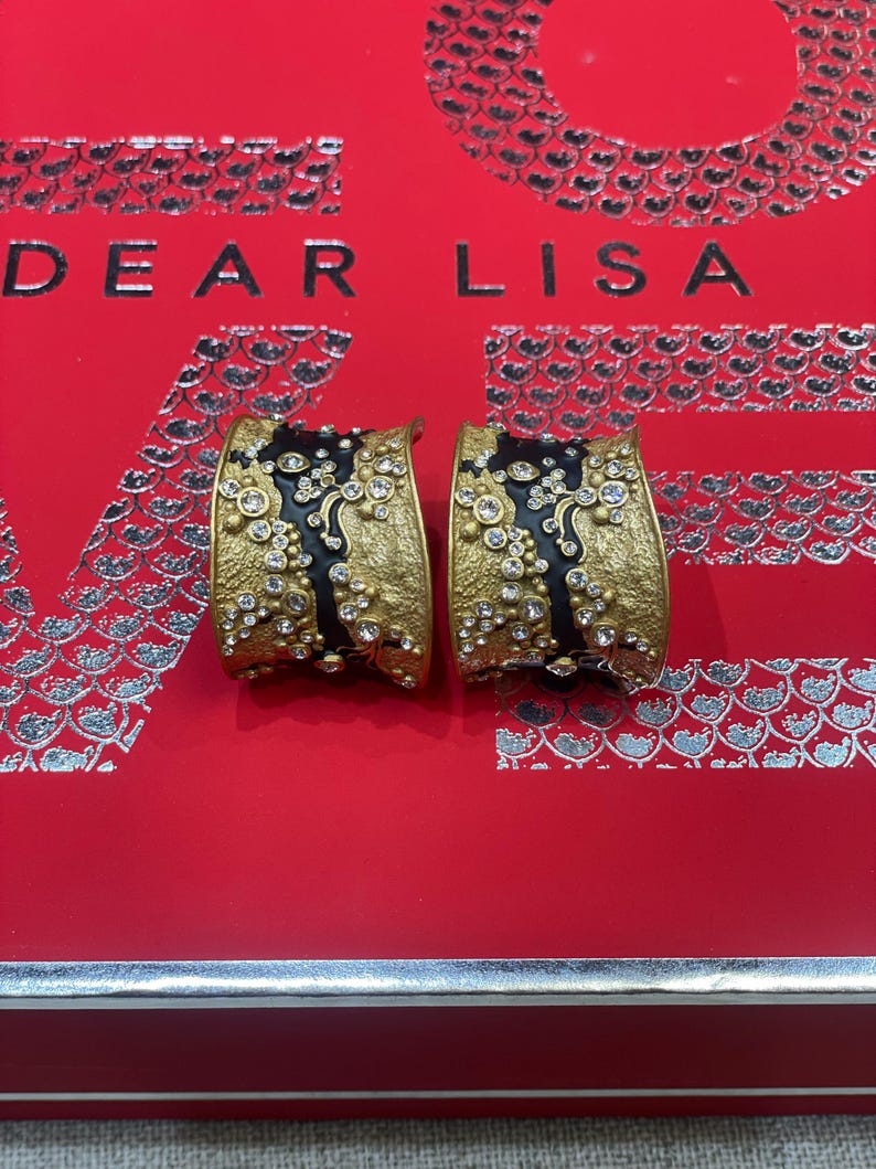May include: A pair of gold-toned earrings with a black enamel center, adorned with clear crystals. The earrings are set against a red background with the text "DEAR LISA". The earrings have a textured finish and a curved design.