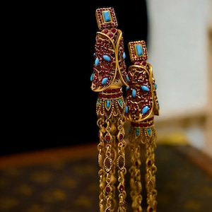 Könnte beinhalten: Ein Paar verzierter goldener Ohrringe. Die Ohrringe haben einen rechteckigen türkisfarbenen Stein an der Spitze, umgeben von roten und türkisfarbenen Akzenten. Lange, baumelnde Ketten mit kleinen goldenen Gliedern und roten Steinen hängen von unten.
