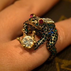 Puede incluir: Un anillo de rana dorado con detalles de gemas verde esmeralda y azul. La rana sostiene una corona dorada con una gema grande y transparente. El anillo tiene gemas rojas para los ojos.